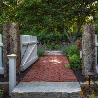 A white garden gate framed by stone posts creates a graceful transition from walkway to garden, blending structure with softness.
