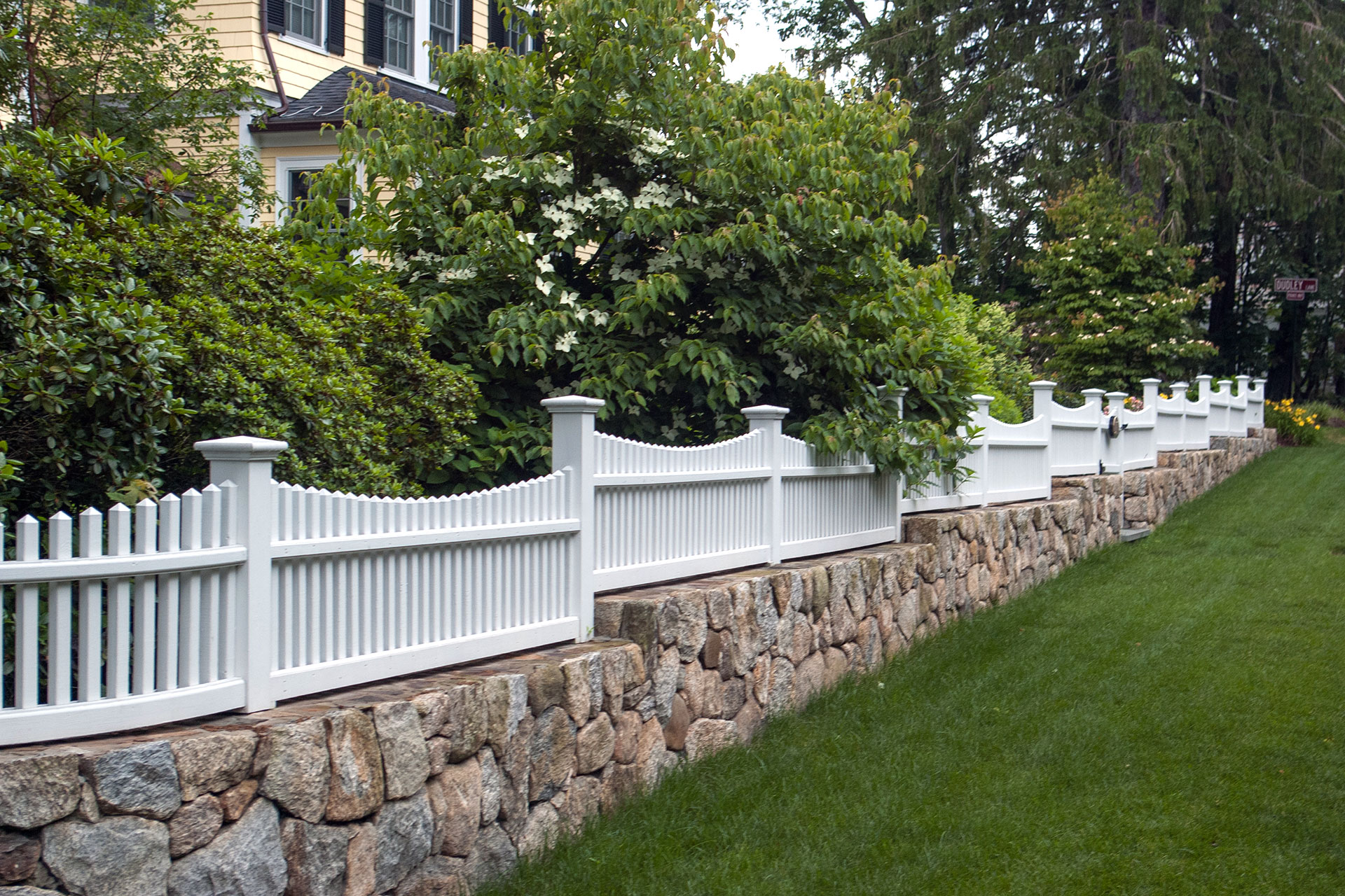 Low-maintenance white vinyl picket fencing complements stone walls in Wellesley, adding curb appeal while clearly marking property lines.