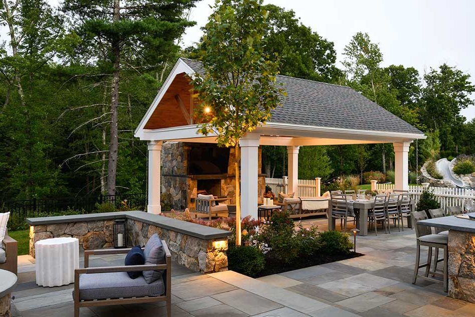 A beautifully crafted garden pavilion anchors this refined outdoor living space, creating a sheltered gathering area on a stone terrace surrounded by layered plantings and soft landscape lighting.