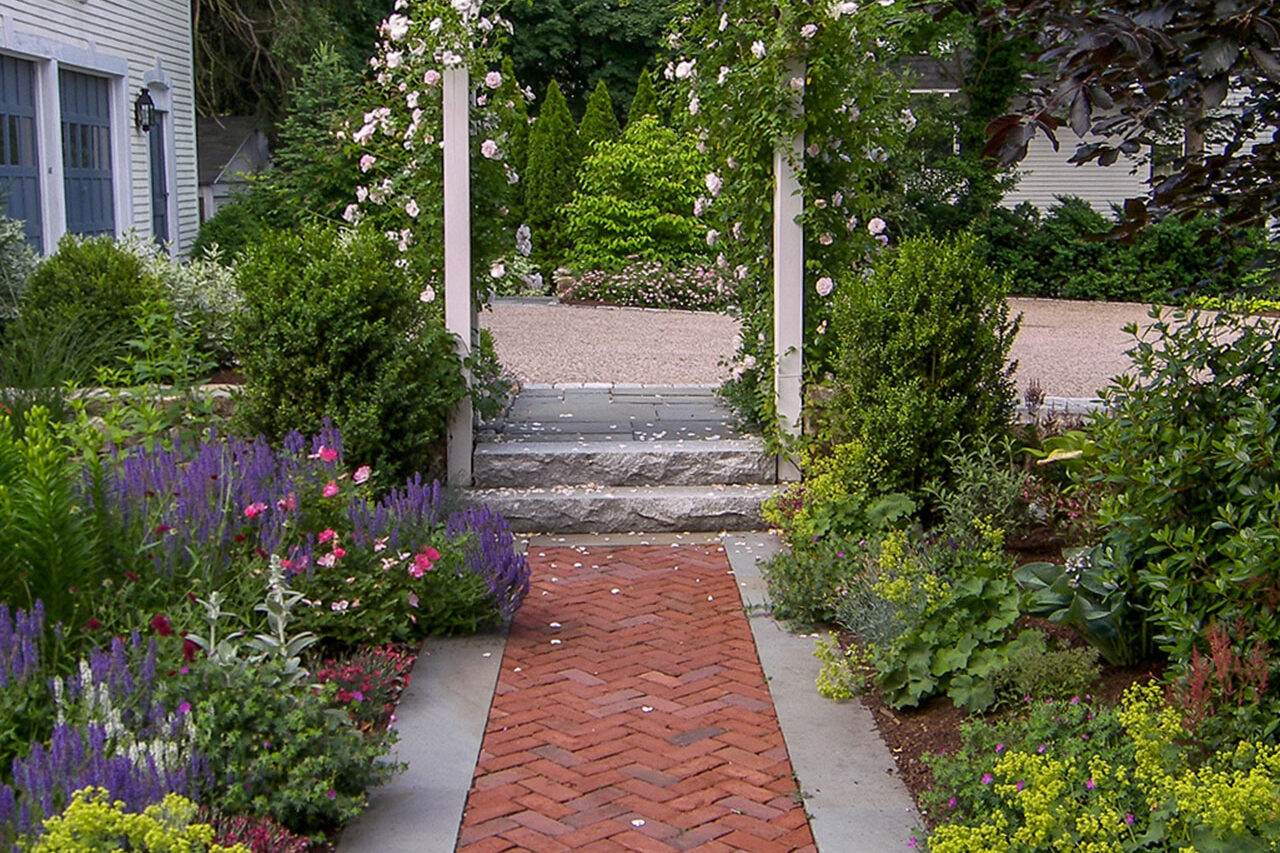 Brick and stone walkway passes through an archway, creating a formal garden entry with architectural presence.