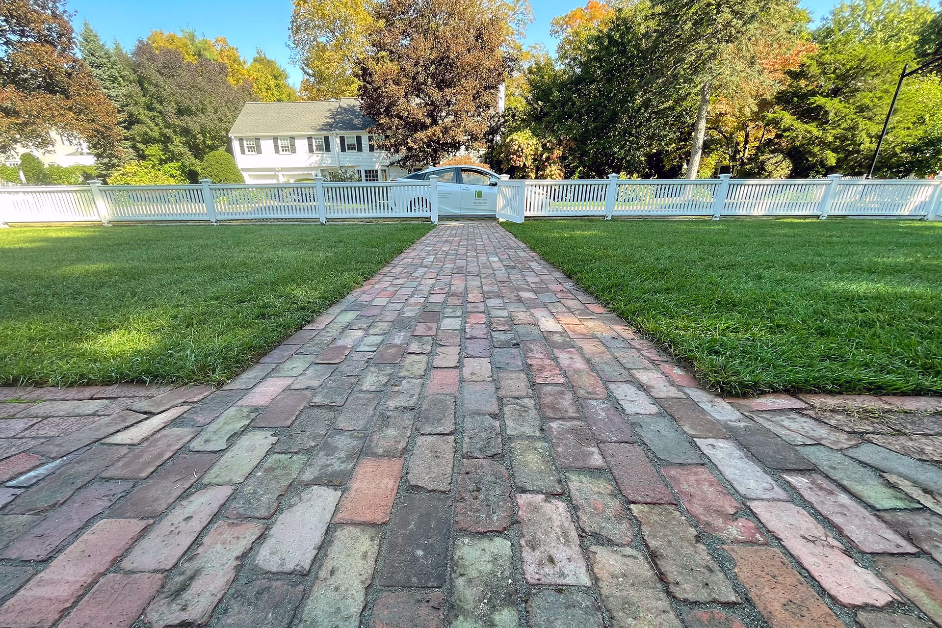 Reclaimed brick walkway leads to a white picket gate, adding texture, warmth, and a welcoming garden transition.