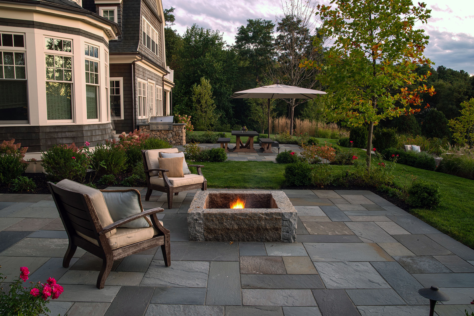 Bluestone patio with built‑in firepit and garden seating offers structured hardscape, cozy fire feature, and lush planting that draws the eye.