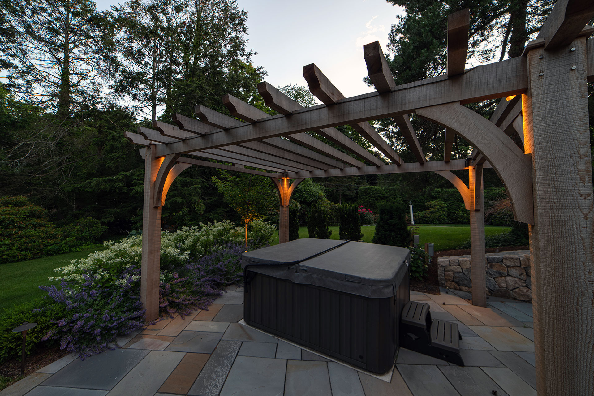 Cedar pergola with integrated lighting elevates a hot tub garden, extending use into the evening while adding privacy and structure.