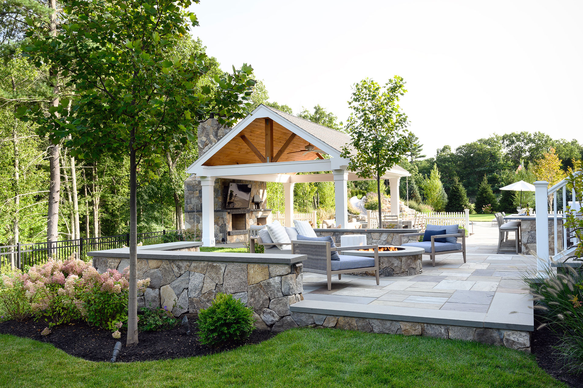 Pergola-style pavilion defines an outdoor dining area with a wood table and grill, improving comfort, flow, and usability on a stone patio.