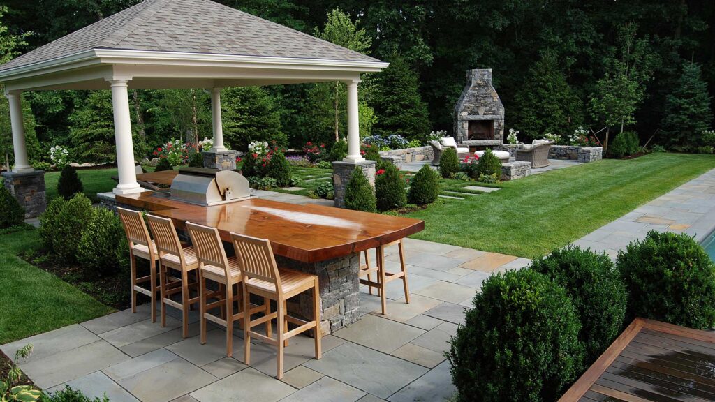 Pavilion-defined dining area on a stone patio pairs a wood table with a built-in grill, adding structure, light shade, and an inviting setting for outdoor entertaining.
