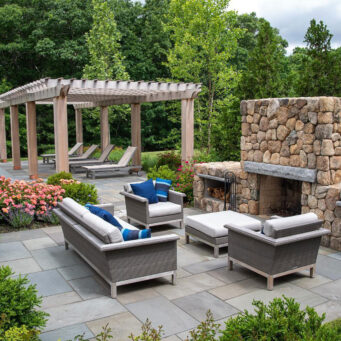 Wood pergola with stone fireplace anchors a poolside living area, enabling multi-season use and dramatic visual impact.