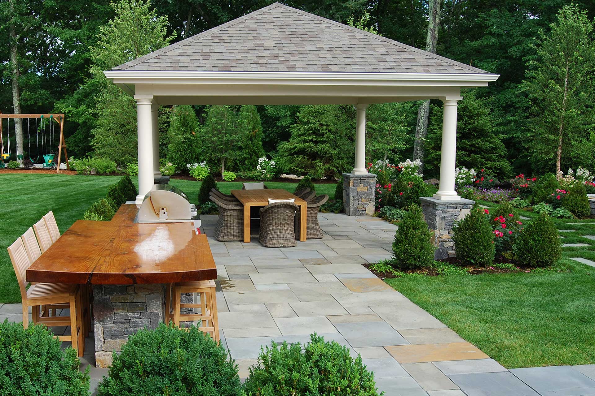 Roofed pavilion anchors a stone patio with dining and outdoor kitchen space, delivering year-round usability and architectural presence.