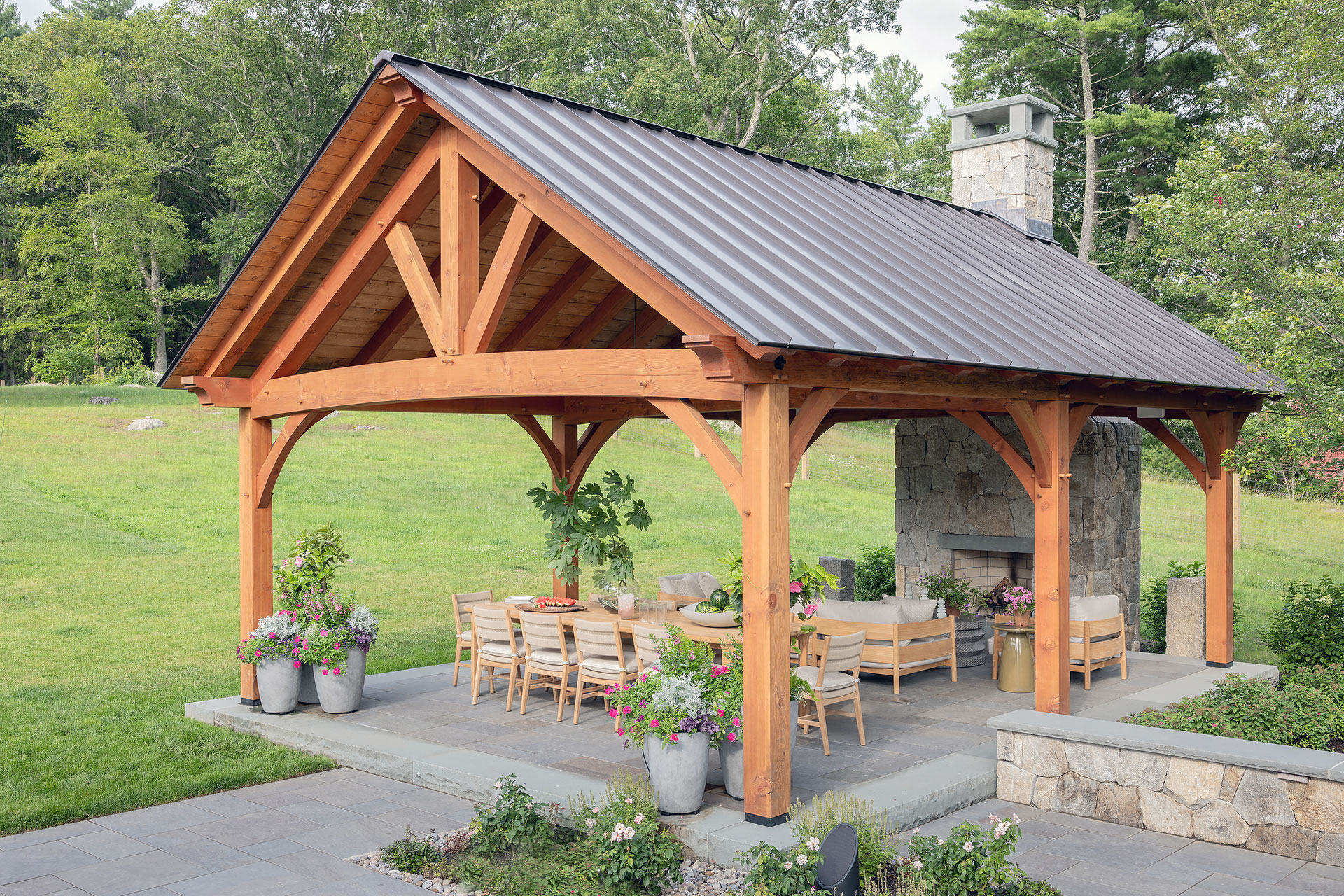 Timber frame pavilion with stone fireplace creates a durable outdoor dining space, offering full shelter, warmth, and year-round usability in a New England landscape.