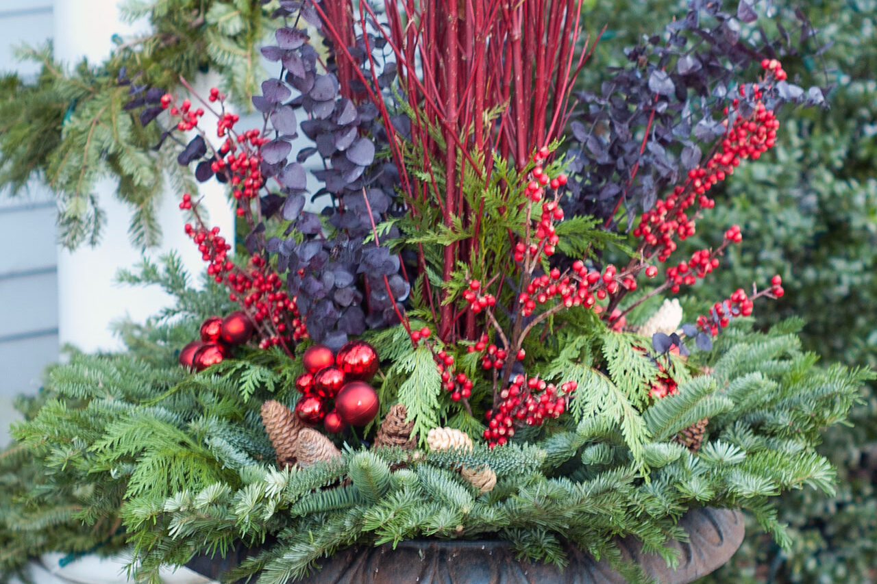 A cast iron urn filled with evergreen boughs and red twig accents creates a festive winter porch display with lasting structure.