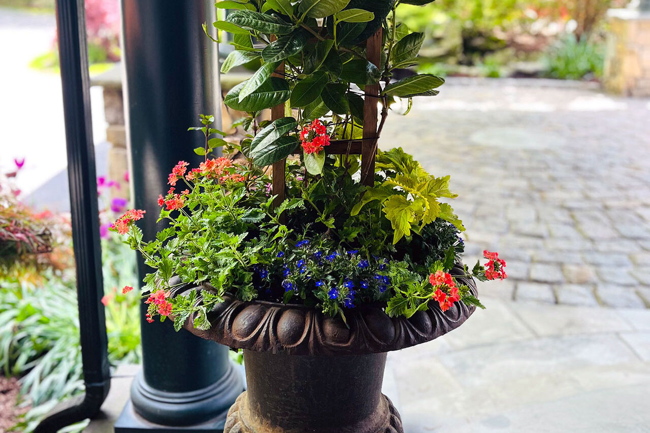 A rusted iron urn on a covered porch features mixed summer annuals, combining durability and warmth while reducing maintenance through sheltered placement.