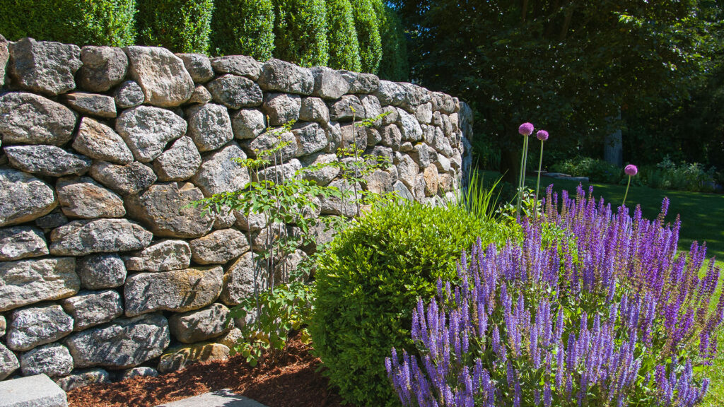 A dry-laid fieldstone retaining wall defines the garden edge, using irregular, hand-set stone to manage a subtle grade change while creating a low, natural boundary with a relaxed, timeless character.