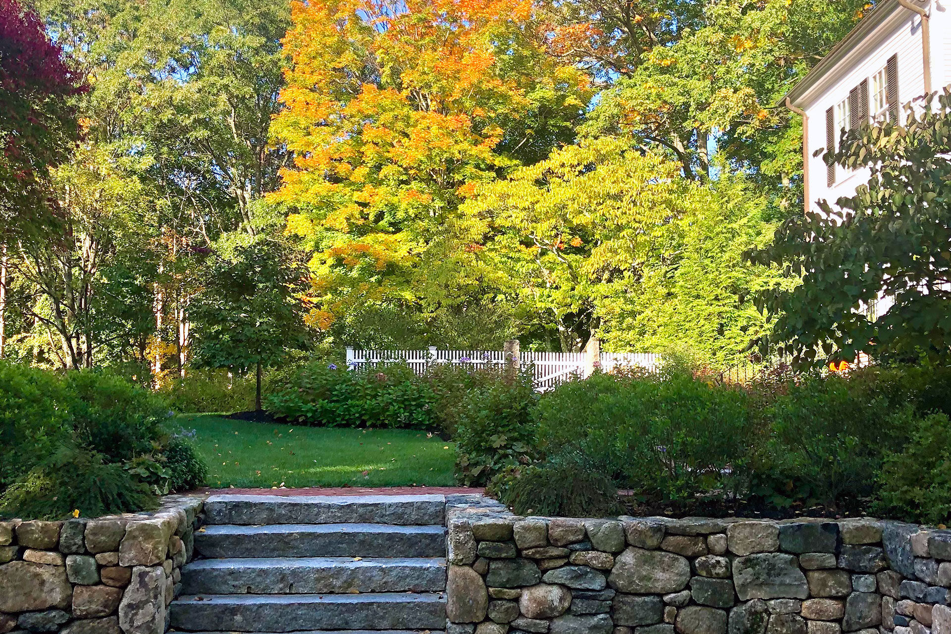 Granite block steps meet a fieldstone wall along the drive approach, with shrubs and hedges helping guide movement while softening the mass of stone.