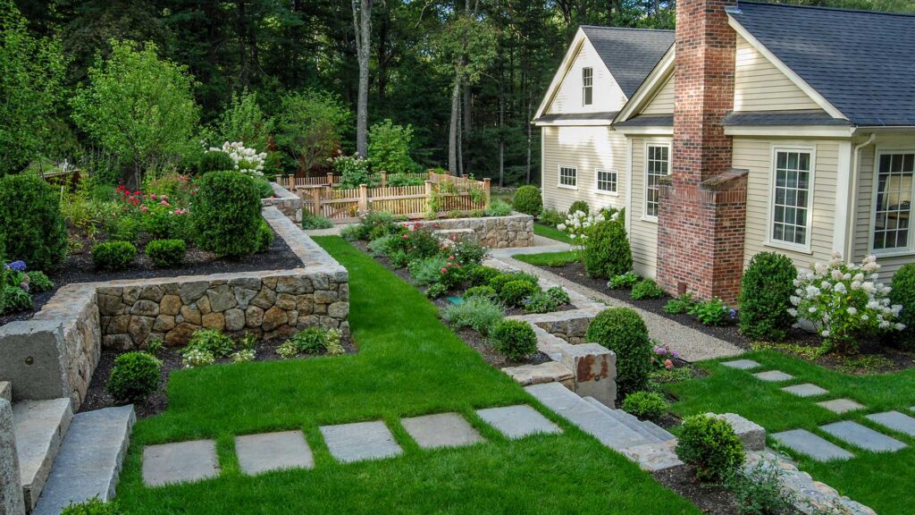 A traditional fieldstone wall anchors the front yard, its weathered texture and warm tones complemented by neatly shaped shrubs and hedges that reinforce symmetry and provide year-round structure near the home.