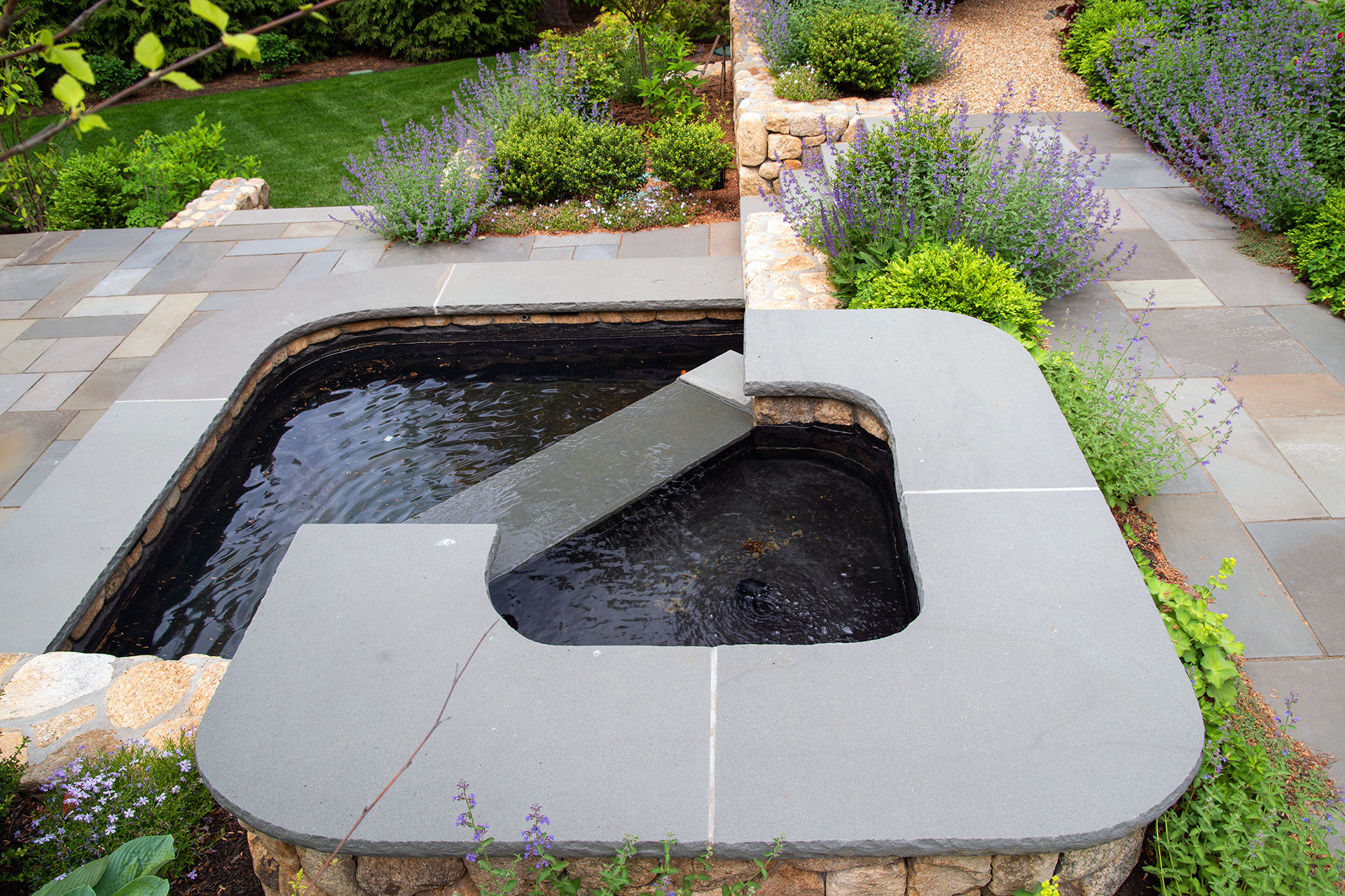 This custom stone wall waterfall channels water through a sculpted granite basin, creating gentle movement and sound while reinforcing clean circulation and a refined focal point within the surrounding patio and planting design.