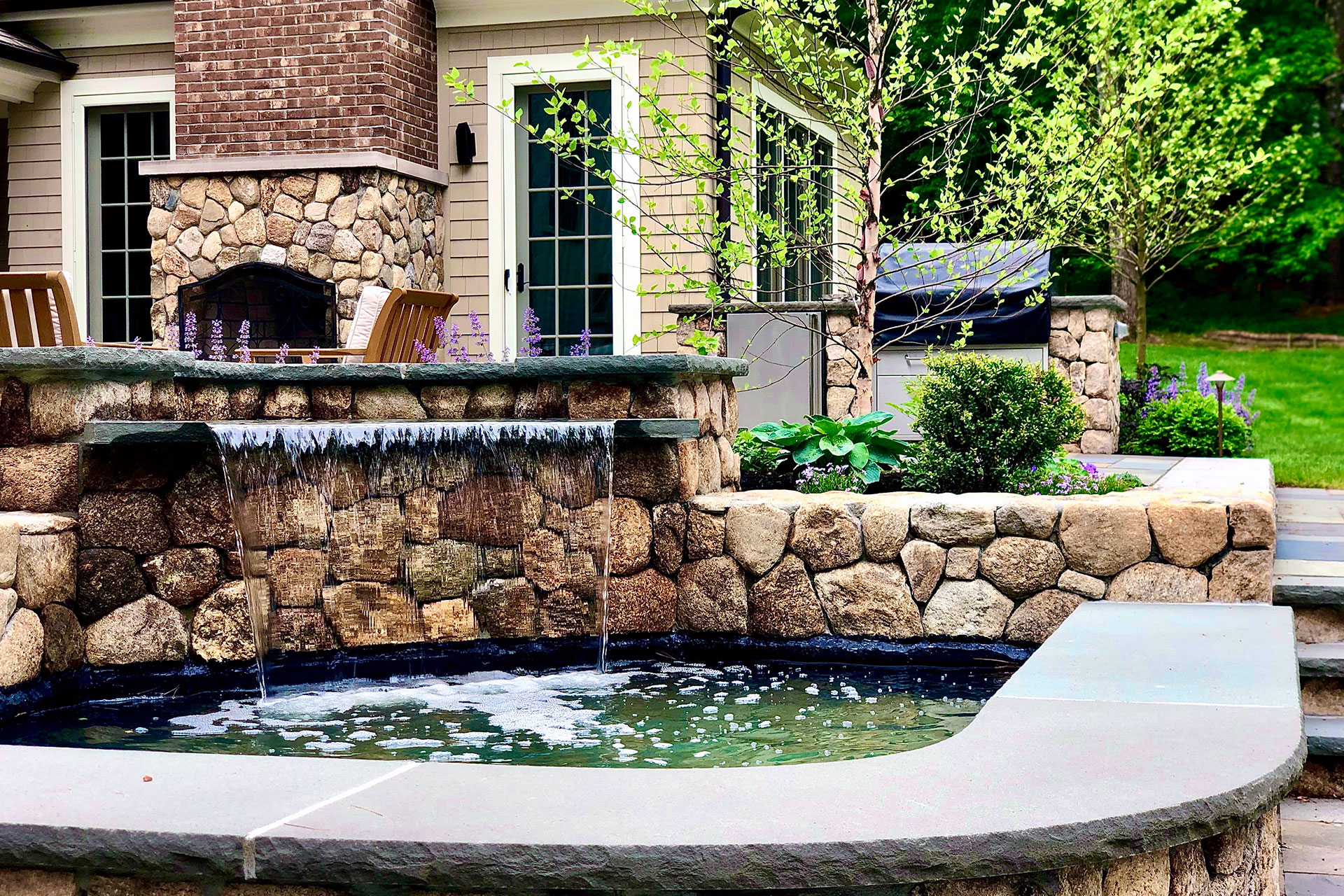 A hand-built stone wall waterfall enhances this patio with gentle sound, visual texture, and a seamless connection between hardscape and surrounding landscape.