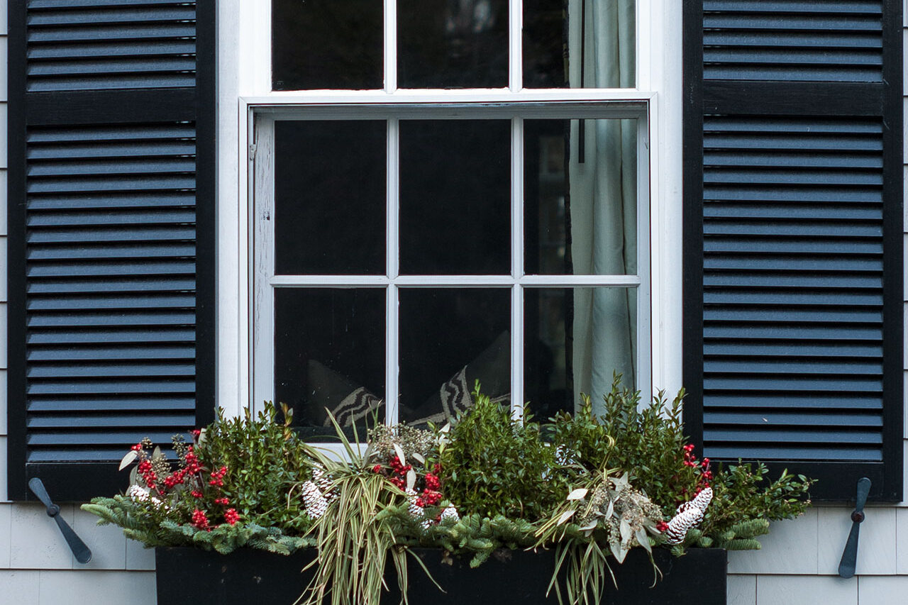 Black metal window box with winter evergreens, red berries, pine cones, and ornamental grasses, adding seasonal structure and curb appeal to a classic New England façade.